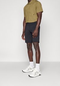 Oliwkowa koszulka polo z kieszonką na piersi, w połączeniu z czarnymi szortami i białymi trampkami z szarymi akcentami oraz białymi skarpetkami.