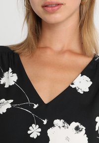 Tessuto nero con stampa floreale bianca, caratterizzato da un design a scollo a V e dettagli plissettati. Texture liscia, focalizzata sui dettagli del décolleté e della parte superiore del petto.
