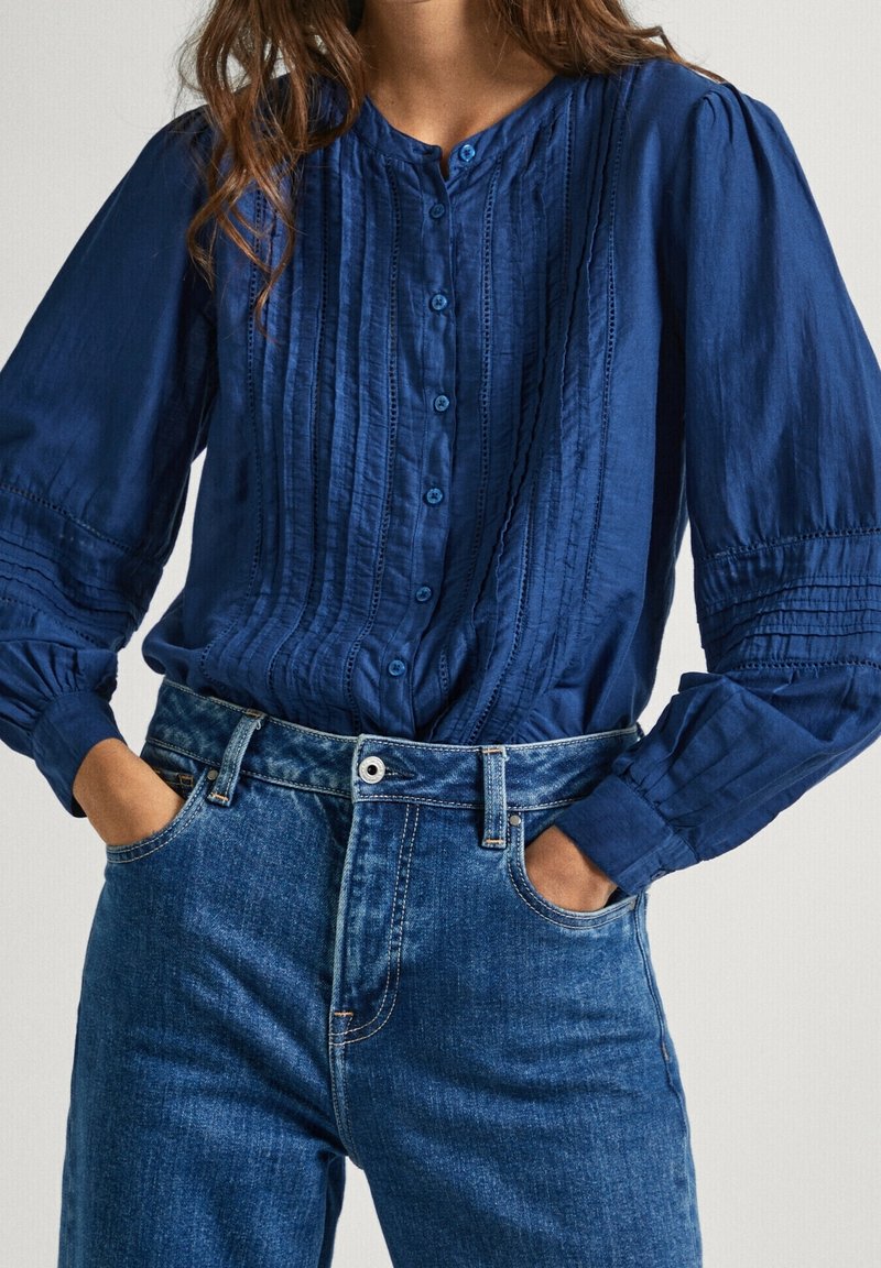 Chemise boutonnée bleu marine avec des rayures texturées, manches longues bouffantes et boutons bleus, associée à un jean en denim taille haute.