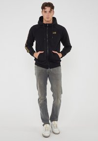 Felpa nera con cerniera, tasca a marsupio, cappuccio con lacci, logo dorato sul petto e scritte sulle maniche. Indossata con jeans grigi sbiaditi e sneaker bianche.