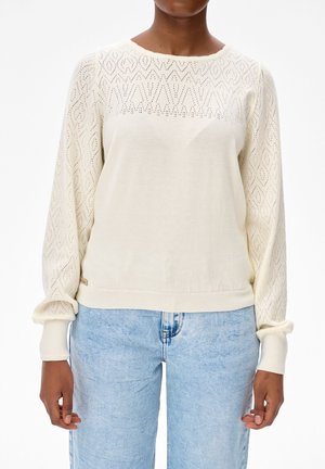 Vrouw draagt een crèmekleurige gebreide trui met diamanten patroon op de mouwen en lichtblauwe high-waist jeans, staand tegen een witte achtergrond.