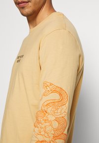 Langärmliges, hellgelbes T-Shirt mit orangefarbenem Schlangen- und Blumenstickerei auf dem linken Ärmel. Weicher Stoff, Rundhalsausschnitt-Design.