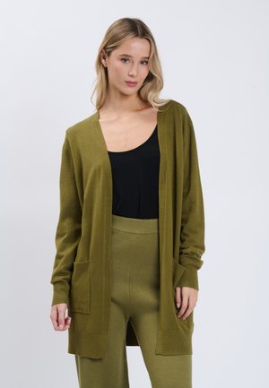Kvinde med blondt hår iført olivengrøn cardigan og bukser over en sort top, stående mod en ensfarvet hvid baggrund.