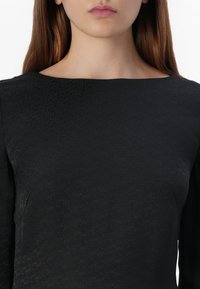 Close-up of a person’s neckline and face wearing a black textured top, with a neutral expression and long brown hair.