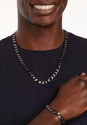 Kettenhalskette und Armband aus ineinandergreifenden schwarzen Metallgliedern. Armband am Handgelenk getragen, Halskette liegt auf dem Kragen eines dunklen T-Shirts.