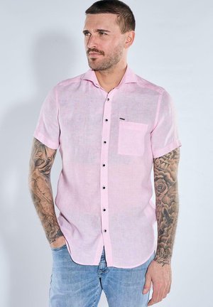 Mann mit kurzen Haaren und Tattoos an beiden Armen, der ein hellrosa kurzärmeliges Hemd und blaue Jeans trägt und zur Seite schaut.
