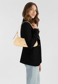 Beige leren handtas met een gebogen silhouette en een verstelbare band, met een subtiel logo. Draag onder de arm met een zwarte blazer.