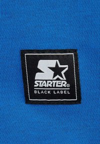 Tessuto di cotone blu con un'etichetta nera prominente che presenta il testo "STARTER" e una grafica a stella, bordata da cuciture bianche.