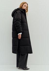 Schwarzer Steppmantel mit Kapuze, mit gestepptem Design, Druckknöpfen und knöchellanger Länge. Kombiniert mit grauen Hosen und Absatzschuhen.