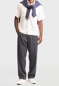 Hombre con camisa de manga corta blanca, suéter de punto gris sobre los hombros, pantalones sueltos gris oscuro y zapatillas blancas, de pie con las manos en los bolsillos.