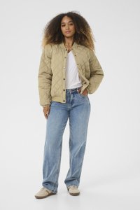 Jachetă bomber bej matlasată cu nasturi tip snap, mansete ribbate și o croială lejeră, combinată cu blugi largi de culoare albastru deschis și adidași bej.