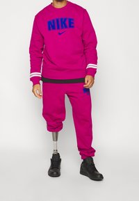 Conjunto deportivo de sudadera y pantalones en fucsia de Nike con logotipo azul en contraste. Presenta puños y cintura acanalados. Zapatilla negra en el pie derecho.