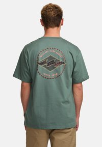 Grünes Baumwoll-T-Shirt mit einem runden Rücken-Graphic, das ein Wellenmotiv und den Text "Qualität Surfartikel seit 1973" zeigt. Mit kurzen Ärmeln.