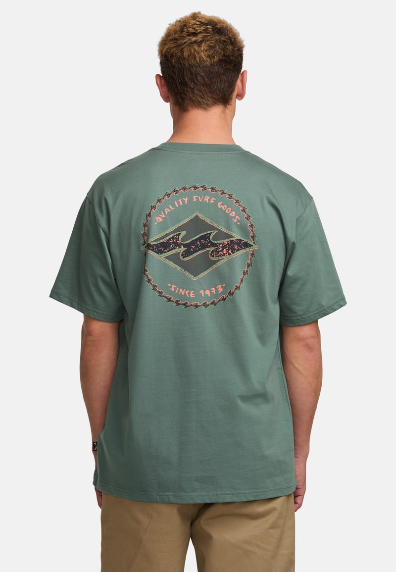 Grünes Baumwoll-T-Shirt mit einem runden Rücken-Graphic, das ein Wellenmotiv und den Text "Qualität Surfartikel seit 1973" zeigt. Mit kurzen Ärmeln.
