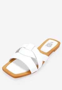 Sandale blanche plate avec des brides croisées, un bout carré et une semelle en caoutchouc beige. Présente un logo discret sur la semelle intérieure.