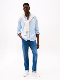 Camisa risca azul claro com um suéter branco drapeado nos ombros, combinada com jeans azuis e ténis brancos. Fundo simples.