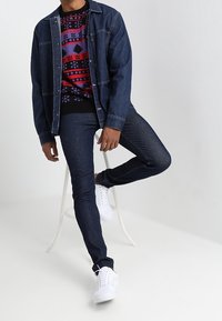 Jeansjacka över en flerfärgad mönstrad tröja, parat med mörka jeans och vita sneakers. Funktioner inkluderar knäppningar och ribbade ärmslut.