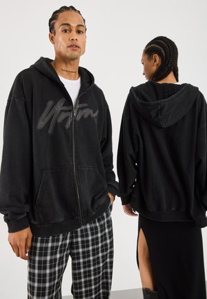 Man facing front wears black zip-up hoodie with large logo and plaid pants; woman facing away wears black hoodie and skirt with high slit.