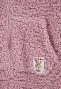 Gros plan sur un tissu rose duveteux avec une fermeture éclair et un patch montrant un ours en peluche et le texte "LET'S GET cozy".
