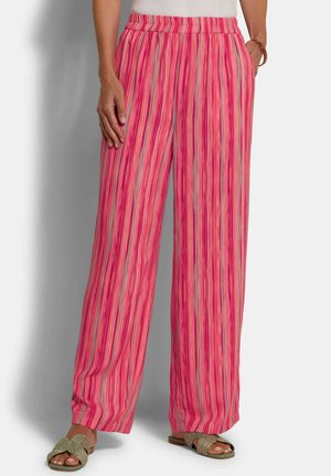 Vrouw die losse roze gestreepte broek draagt met een elastische tailleband, staand met één hand in de zak en groene gevlochten sandalen.