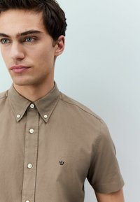 Camisa de botones de manga corta en color beige, de algodón, con un logotipo de corona negra en el pecho y botones blancos a lo largo de la tapeta. Textura suave.