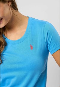 Lys blå t-skjorte med korte ermer laget av mykt stoff, med rund hals og en liten rød brodert logo på brystet.