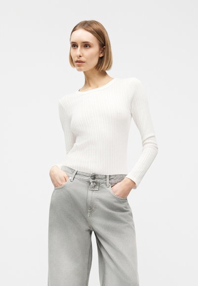 Γυναίκα με κοντά ξανθά μαλλιά, που φορά λευκό ριμπ μπλουζάκι μακρυμάνικο και χαλαρό γκρι τζιν, με τα χέρια στις τσέπες, κοιτάζοντας ελαφρώς πλάγια.