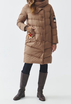 Frau trägt einen knielangen braunen Steppmantel mit Teddybären-Applikationen auf Taschen und Ärmel, schwarze Hosen und braune Stiefel.