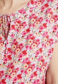 Blusa floral con una tela ligera decorada con flores en tonos rosa, rojo y lavanda. Tiene un escote redondeado y un detalle de lazo en la parte frontal.