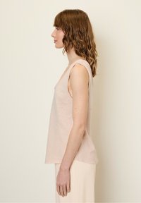 Jasnoróżowa bezrękawnik o luźnym kroju, z surowymi brzegami i subtelną teksturą. Noszony z lekko dopasowanymi jasnymi spodniami.