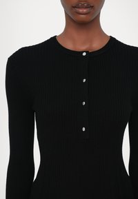 Černý žebrovaný top s dlouhými rukávy, kulatým výstřihem a pěti dekoračními knoflíky vpředu. Hladký materiál a přiléhavý střih.