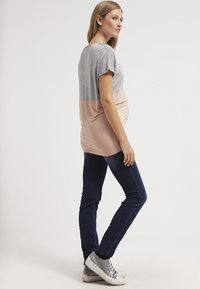 T-shirt de maternidade em cinza e design em blocos de cor rosa claro, com mangas curtas e corte descontraído. Combinado com jeans skinny azul escuro e ténis cinzentos.