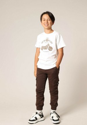 T-shirt graphique blanc avec impression de moto marron, pantalon cargo marron avec poignets élastiques, et baskets noires et blanches.