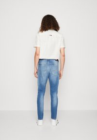 Camisa blanca recortada con patrón de cuadritos, combinada con jeans de mezclilla azul claro, mostrando los bolsillos traseros y zapatillas blancas, sobre un fondo liso.