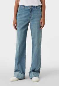 Jasnoniebieskie jeansy z denimu o szerokich nogawkach, z klasycznym pięciokieszeniowym krojem, subtelnym blaknięciem i podwiniętymi mankietami, zestawione z białym obuwiem.