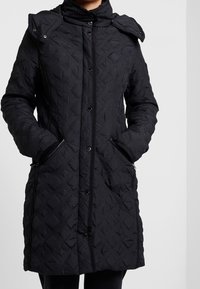 Černá prošívaná bunda s vysokým límcem, patentními knoflíky, zipovými kapsami a texturovanými diamantovými vzory na látce.