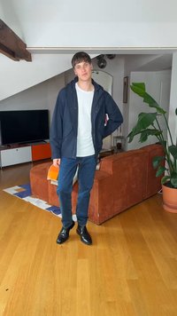 Nuori mies seisoo olohuoneessa yllään laivastonsininen takki, valkoinen paita, siniset farkut ja mustat saappaat, käsi vyötäröllä oranssin sohvan ja suuren kasvin lähellä.