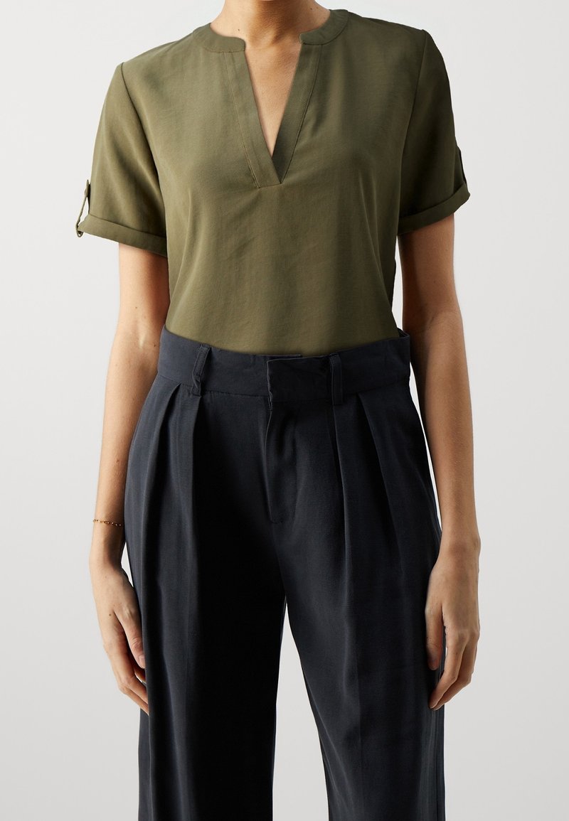 Femme portant un chemisier vert olive à manches courtes avec un col en V, rentré dans un pantalon taille haute plissé bleu marine foncé avec une ceinture.