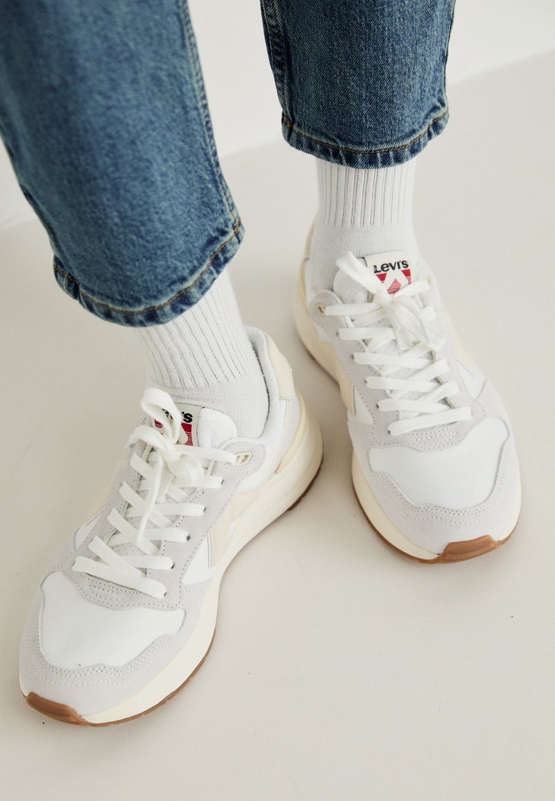 Witte sneakers met beige accenten, veters en een textuur op de teentip. Gecombineerd met geribbelde witte sokken en opgerolde blauwe denim jeans.