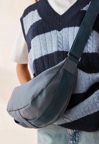 Grå crossbody taske med en glat, mat overflade, der har en bred, vævet teal-strop og en subtil buet form.