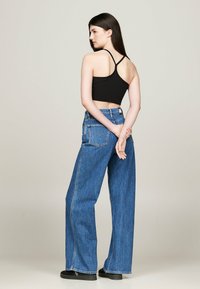 Donna con lunghi capelli scuri, che indossa un crop top nero a spalle scoperte, jeans blu a gamba larga e scarpe nere, in piedi con le mani dietro la schiena.