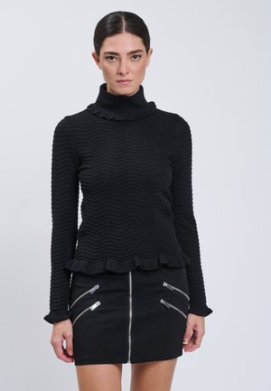 Donna con capelli scuri raccolti indossa una maglia nera a maniche lunghe con texture e una minigonna nera con molteplici cerniere argentate.