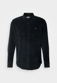 Camisetta in velluto a coste nera a maniche lunghe con colletto botton-down, chiusura frontale con bottoni e sottile logo CK sul petto. Tessuto testurizzato.