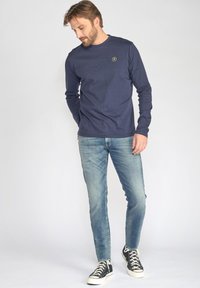 Marineblauw longsleeve shirt met ronde halslijn en klein embleem, gecombineerd met lichtblauwe jeans en zwarte hoge sneakers.