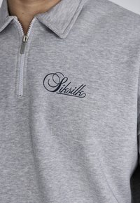 Felpa grigia con zip caratterizzata da un colletto e un logo ricamato blu navy. Texture morbida con un motivo melange.