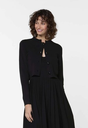 Femme aux cheveux bouclés portant un cardigan noir boutonné par-dessus une robe plissée noire, souriante et regardant sur le côté, sur un fond uni.