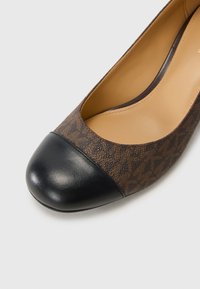 Smeđe cipele sa uzorkom, ravnog kroja, s crnim kožnim kapicom na prstima. Imaju zaobljen oblik i glatku kožnu unutrašnjost. Lagan dizajn.