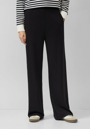 Pantalon noir à jambes larges en tissu doux avec poches latérales, associé à un haut rayé bleu marine et blanc et des chaussures beige sans lacets.