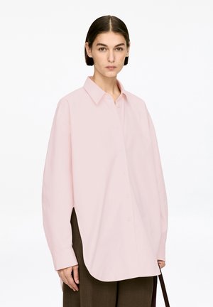 Light pink oversized shirt with a pointed collar, long sleeves, and a curved hem. Features front button closure and side slits.
