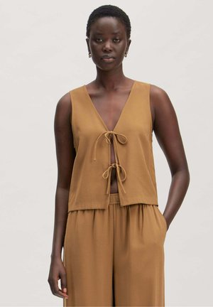 Vrouw draagt een mouwloos beige topje met twee strikjes aan de voorkant en bijpassende wijde broek, staand tegen een effen lichte achtergrond.